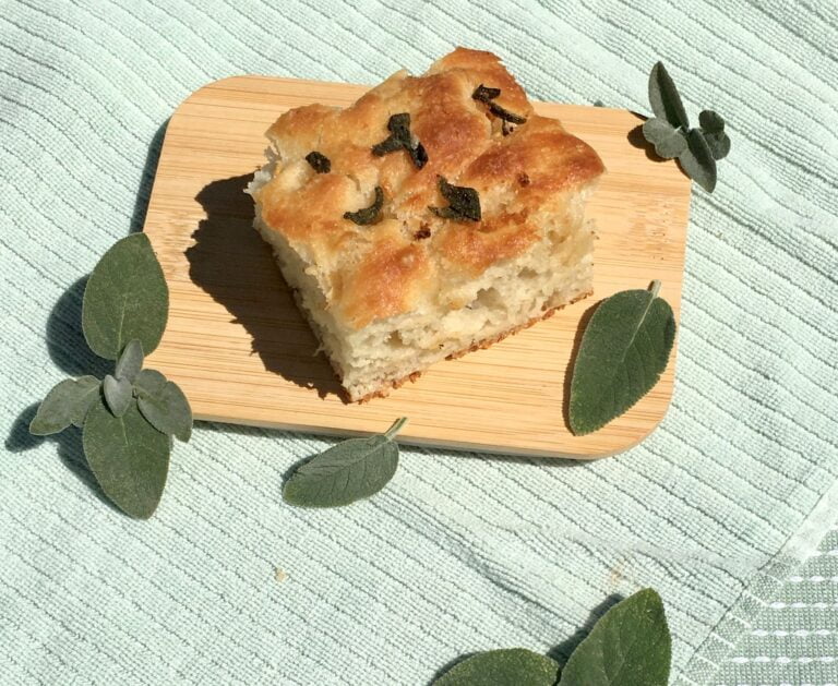 Slice of vegan brown butter and sage focaccia bread. Surrounded by sage leaves.